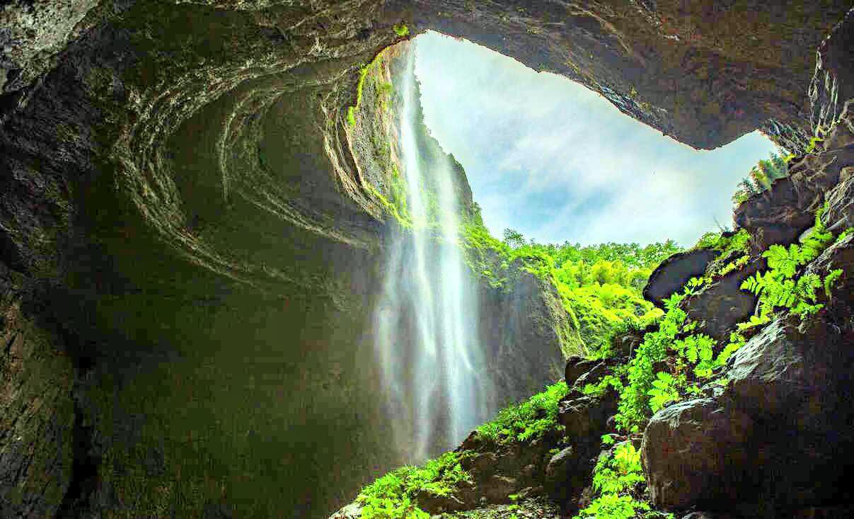 图片展示了从洞穴顶部倾泻而下的瀑布，四周覆盖着绿色植被，透过洞口可见天空。整体景象自然而宁静。