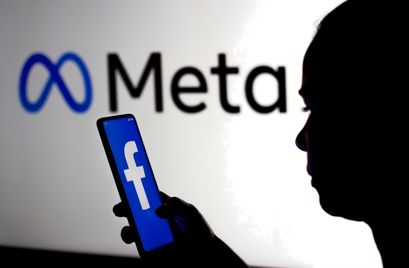 图片展示一位剪影人物手持带有Facebook标志的手机，背景是亮起的“Meta”字样的标识。