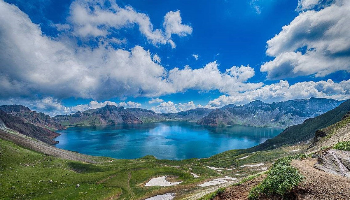 这是一张高山湖泊的照片，湖水呈蓝绿色，周围有草地和雪山，天空中云朵蓬松，景色宁静而壮丽。
