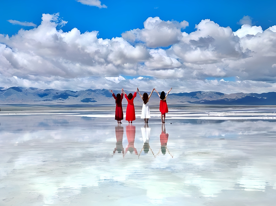 图片展示了四位穿着不同颜色长裙的人在天空和水面映成一色的盐湖上欢快地举手，远处是连绵的山脉。