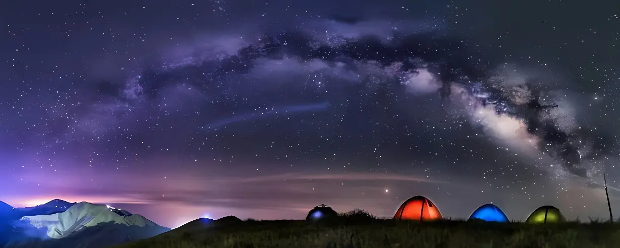 图片展示了壮观的银河横跨夜空，下方是几顶露营帐篷，在山丘上闪烁着彩色的光。