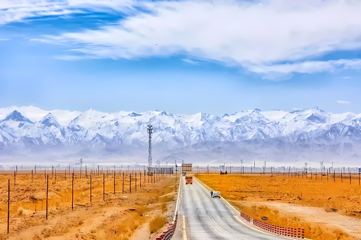 这张图片展示了一条穿越干旱地区通往远方雪山的笔直公路，旁边是电信塔和稀疏的植被，天空呈现出蓝色和白云。