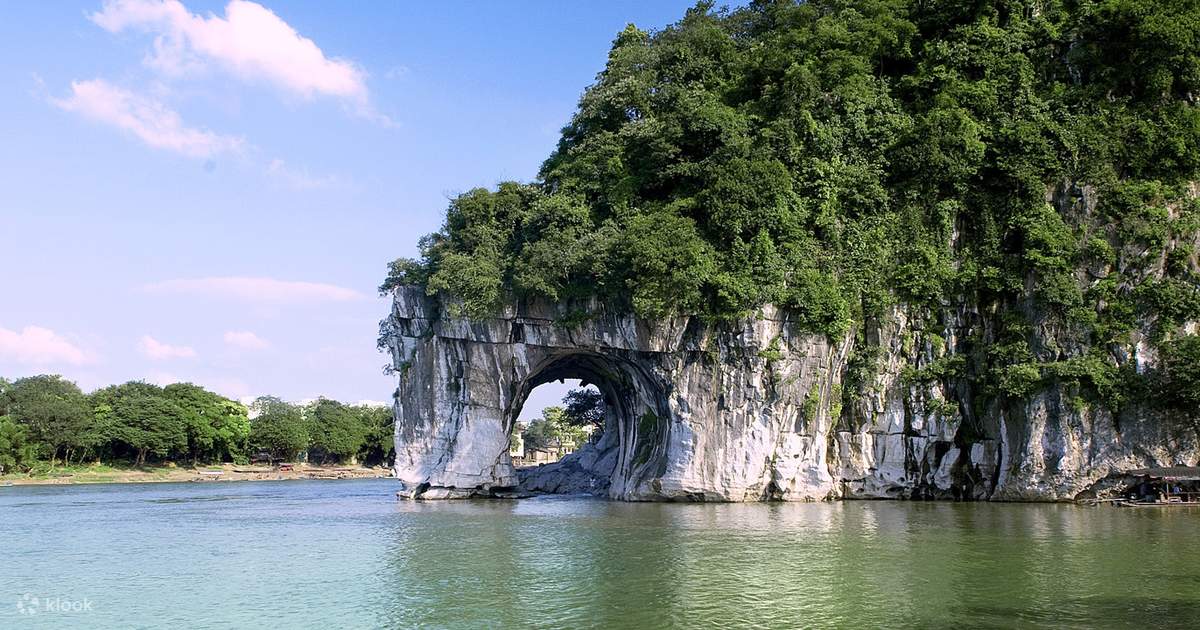 这是一张风景照片，展示了一座巨大的岩石山体，山体中有一个天然形成的大洞，前方是宁静的水面，背后是蓝天和树木。