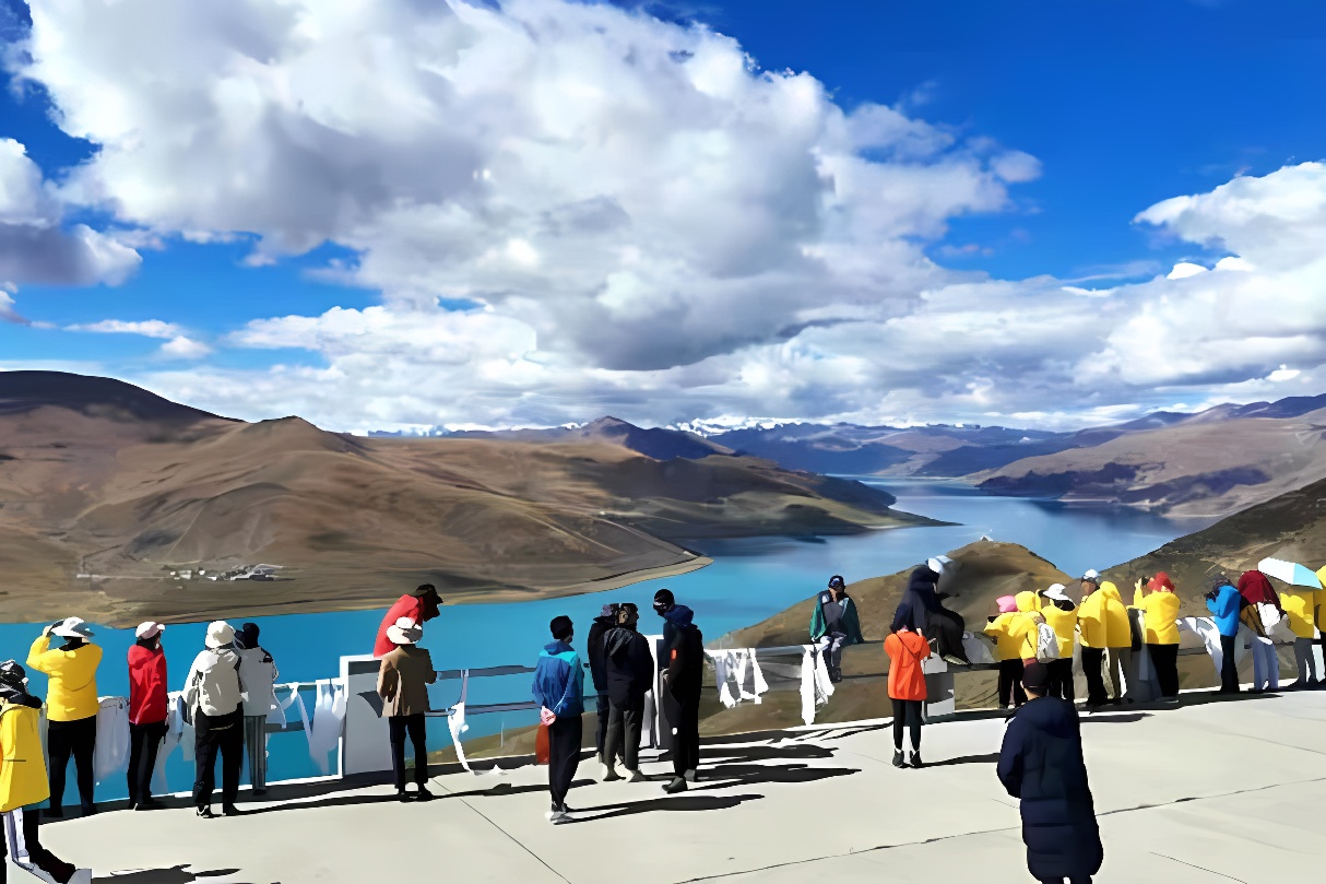 图片展示了一群游客在晴朗天气下观赏山脉间蜿蜒的湖泊，湛蓝的天空下云朵飘浮，景色宁静而壮丽。