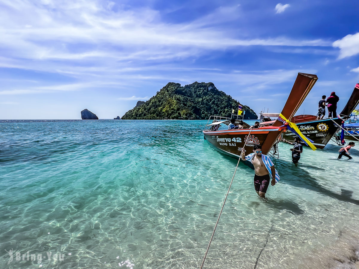 图片展示晴朗天气下，碧绿清澈的海水和沙滩，几艘泰式长尾船停靠在岸边，远处有岛屿。