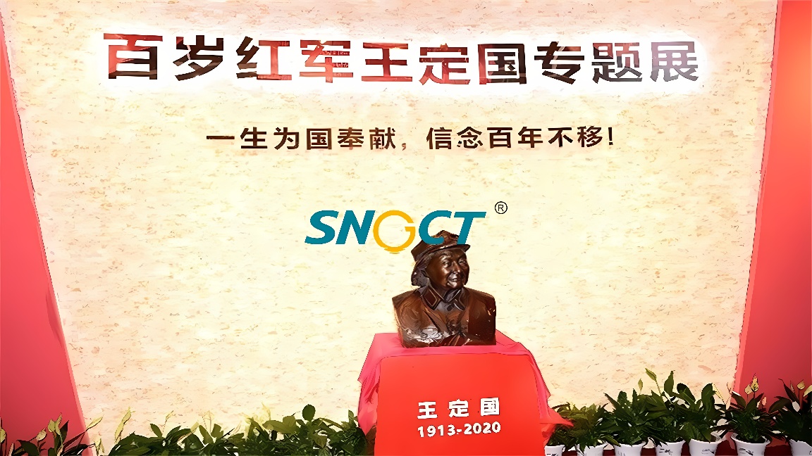 图片展示了一尊铜色半身雕像，前方有介绍文字，背景是红色带有中文标语的墙面，下方有绿色植物装饰。