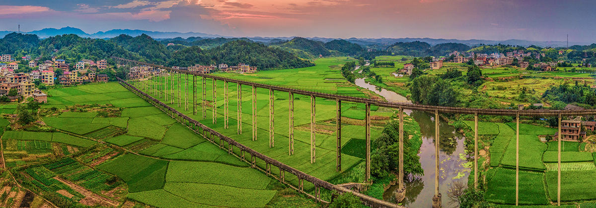 这是一幅田园风光图，展示了一座横跨河流的吊桥，两岸是绿色的稻田和散布的房屋，远处有群山，天空呈现晚霞。