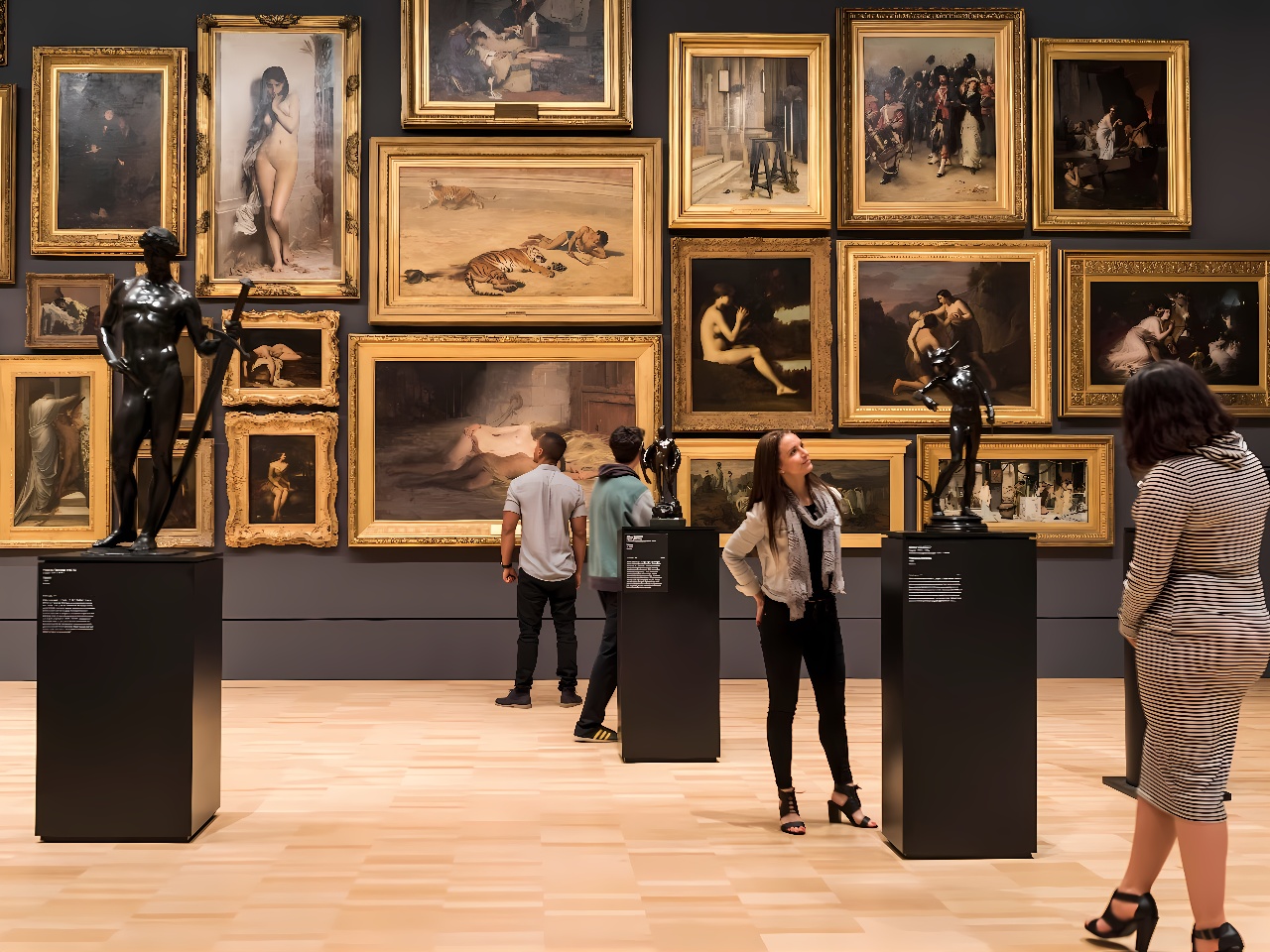 图片展示了几位参观者在艺术画廊内欣赏悬挂在墙上的多幅画作和展示台上的雕塑作品。