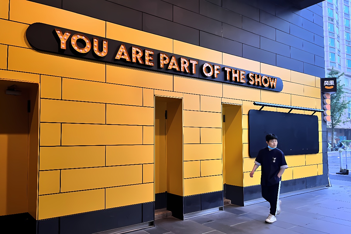 图片展示一位男士从装饰有黄色砖块和“YOU ARE PART OF THE SHOW”字样的墙面旁边的门走出，背景为现代建筑。