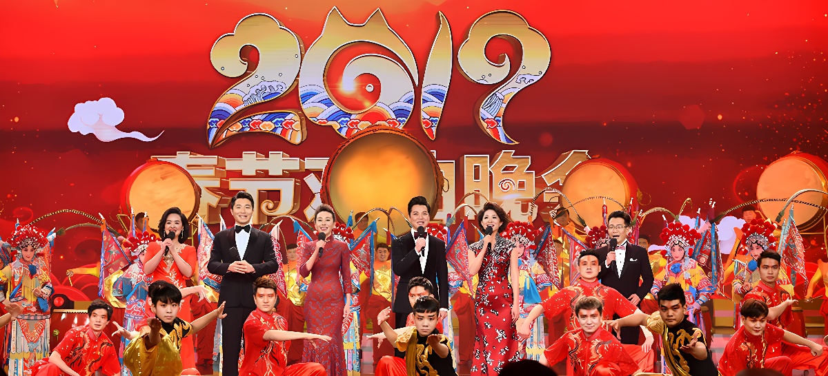 图片展示了一台红色背景的舞台表演，多位身着节日传统服装的演员正表演节目，气氛喜庆，色彩丰富。