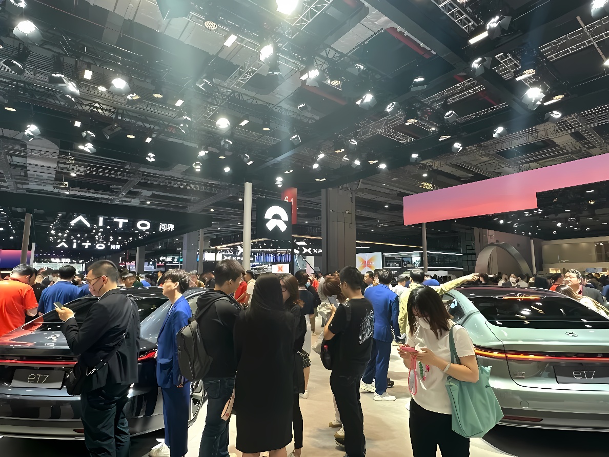 图片展示了一场车展，人群聚集，观看展出的多款汽车。展厅内布置现代，品牌标识清晰可见。