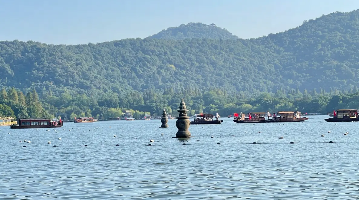 图片展示了一片宁静的湖面，湖中央有一座石塔，远处是青翠的山峦，湖上有几艘船只。