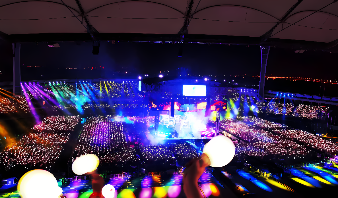 这张图片显示了一个室内体育场内的音乐会现场，舞台上灯光闪烁，观众区手持荧光棒，营造出热烈的气氛。