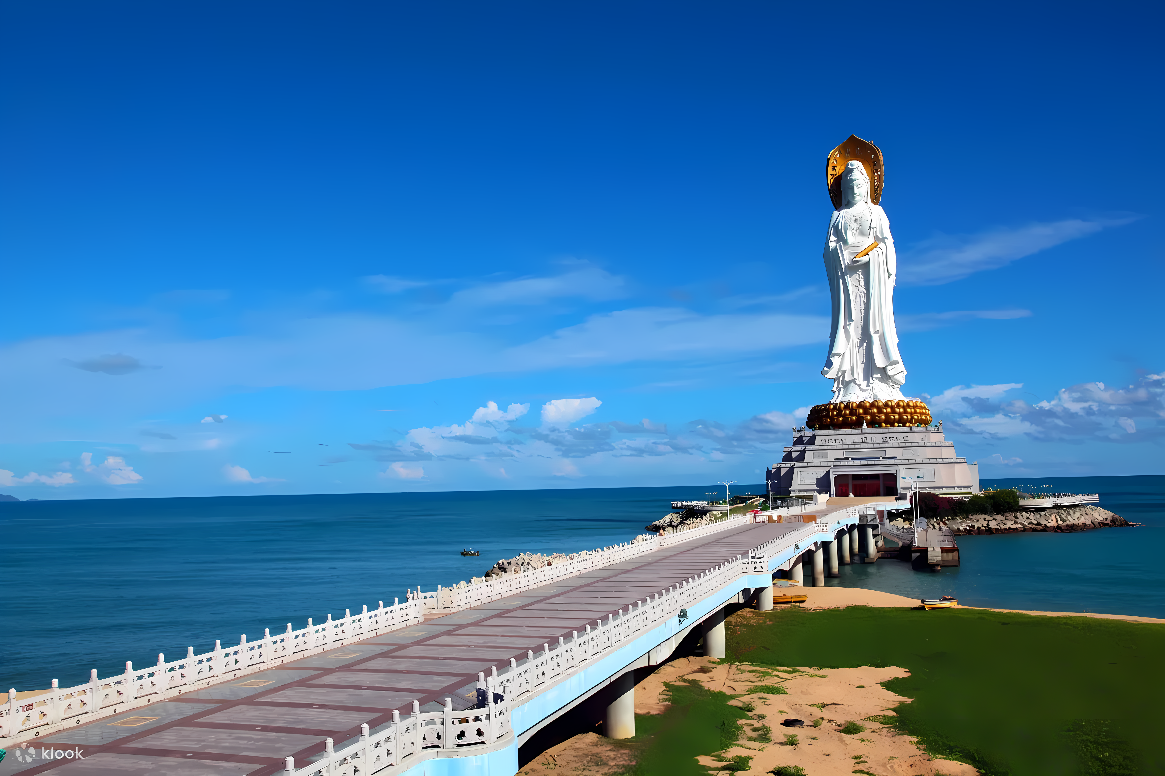 图片展示了一尊巨大的白色观音雕像，位于海边，下方有一座长桥通往雕像，背景是晴朗的蓝天和海洋。