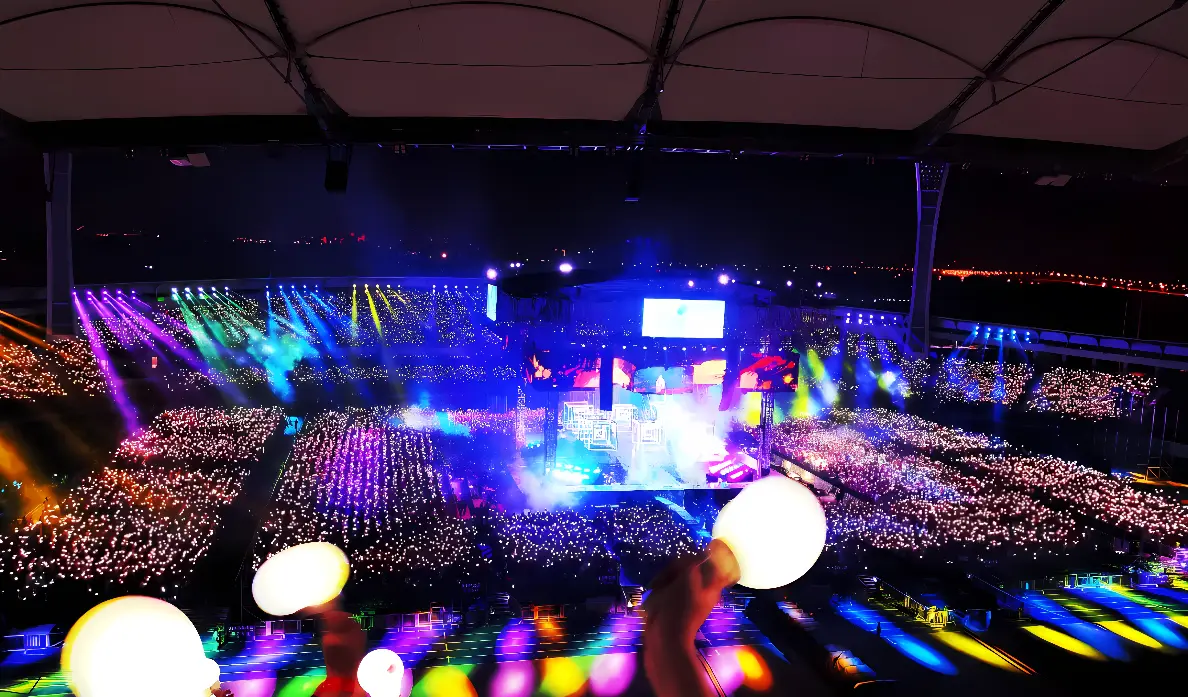 这张图片展示了一场室内演唱会，现场灯光绚烂，人群中举着荧光棒，舞台中央有巨型屏幕，气氛热烈。