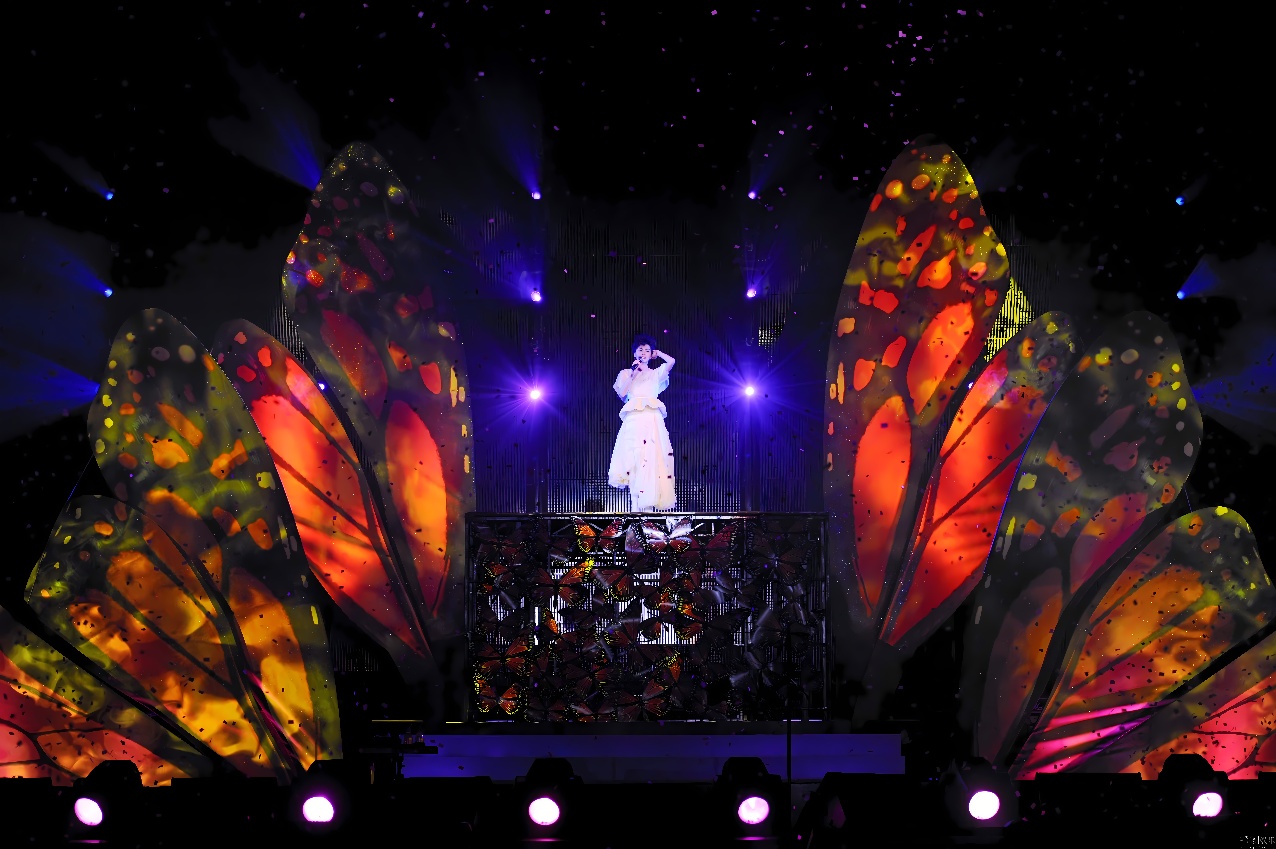 图片展示了一位穿着白色礼服的人站在舞台上，背后是巨大的彩色蝴蝶翅膀装置，舞台灯光照耀下，场面壮观。