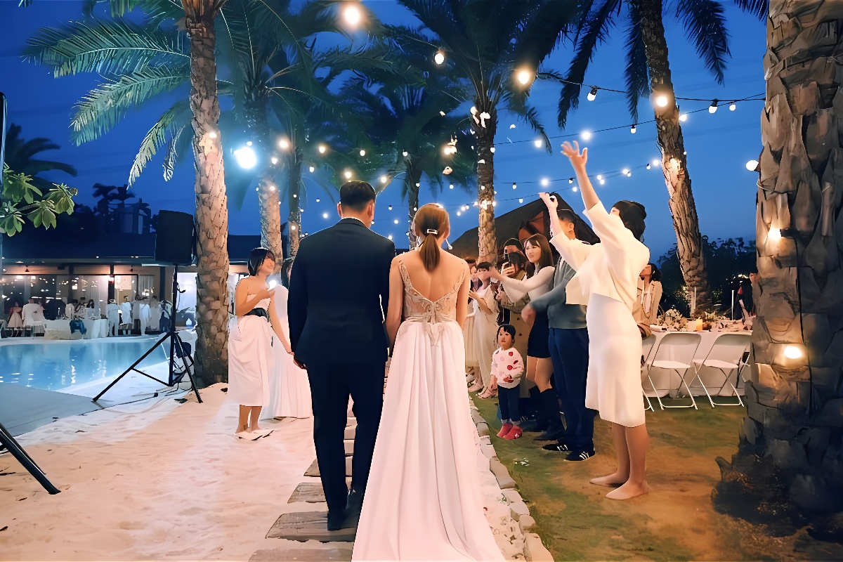 图片展示一对新人在夜幕下的户外婚礼上，走过铺满沙子的小径，两旁是祝福的宾客和璀璨的灯光装饰。