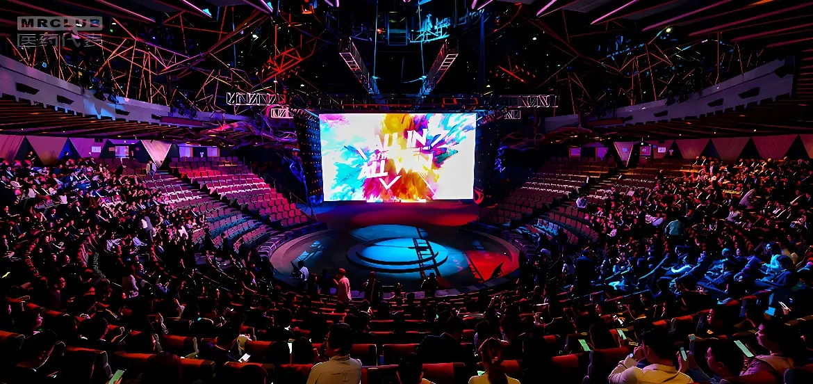 这是一张室内演出场馆的照片，观众席半满，中央有大屏幕，舞台照明五彩斑斓，氛围活跃。