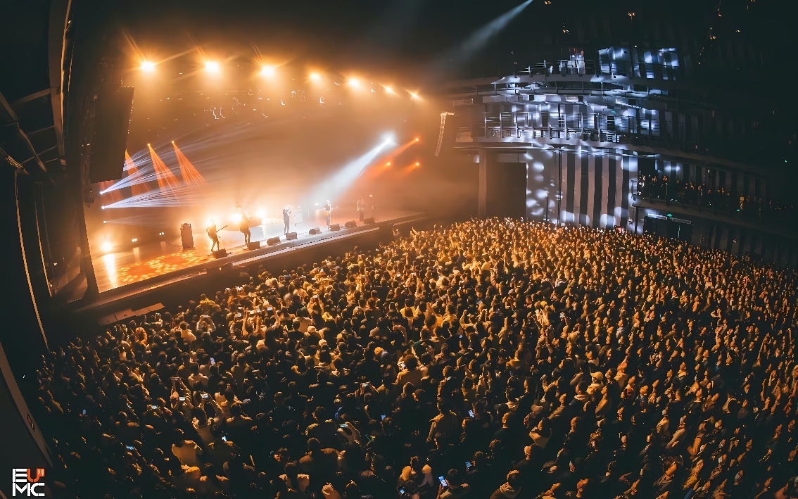 图片展示了一个室内音乐会现场，舞台上灯光闪烁，乐队正在表演，下方观众区密集的人群在享受音乐和演出。