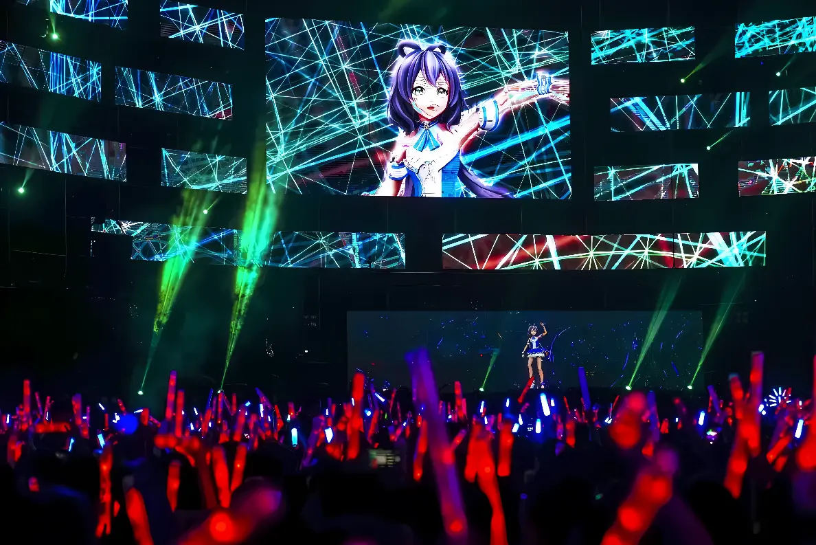 图片展示了一场虚拟偶像音乐会，舞台上大屏幕显示着三维动漫角色，下方观众手持荧光棒，氛围热烈。