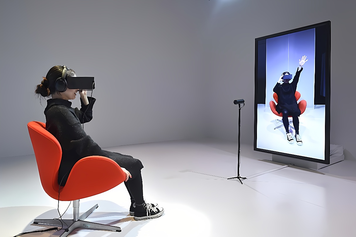 图片展示一位女性坐在橘红色椅子上，戴着虚拟现实头盔，面前是显示她虚拟形象的大屏幕。