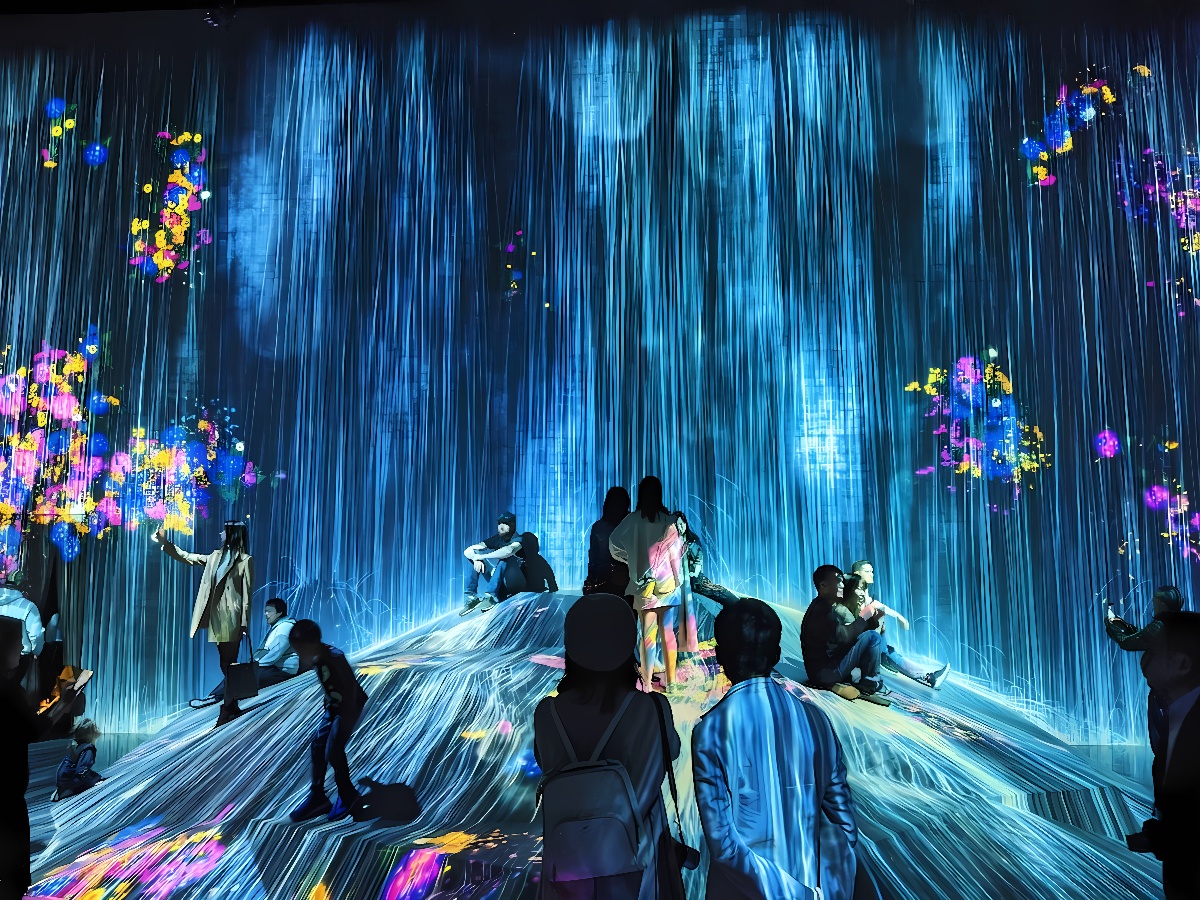 图片展示了一群人在充满光影效果的展览中观赏，仿佛置身于虚拟的瀑布和五彩斑斓的花朵之中。
