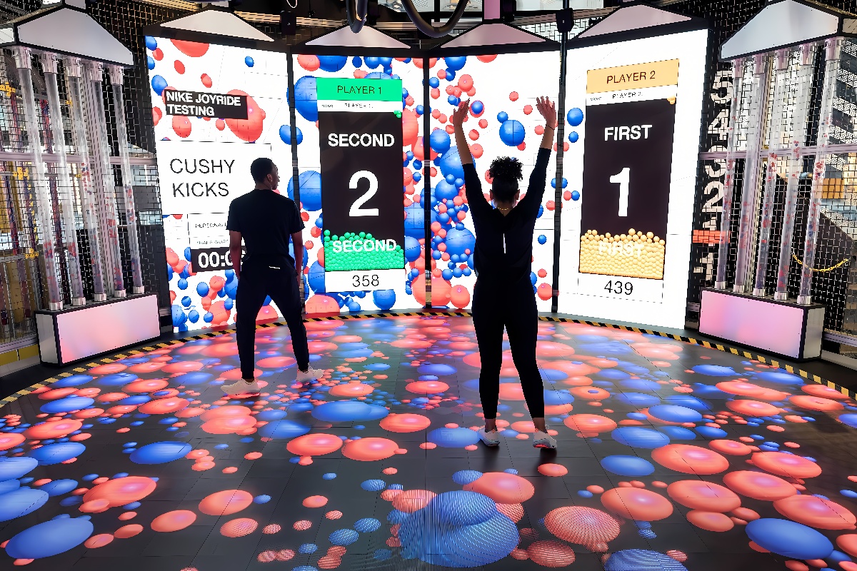 图片展示两人在互动式屏幕前玩游戏，地面是虚拟互动界面，四周装饰现代化，氛围活跃。