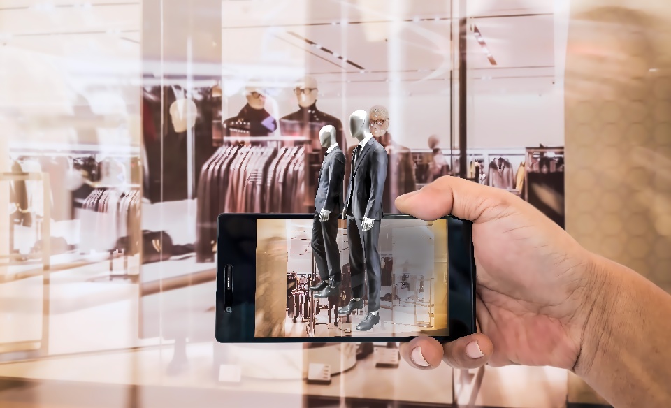 一只手拿着手机对着橱窗拍照，手机屏幕显示橱窗内的场景，包括穿着时尚服装的人体模特和背后的衣物。