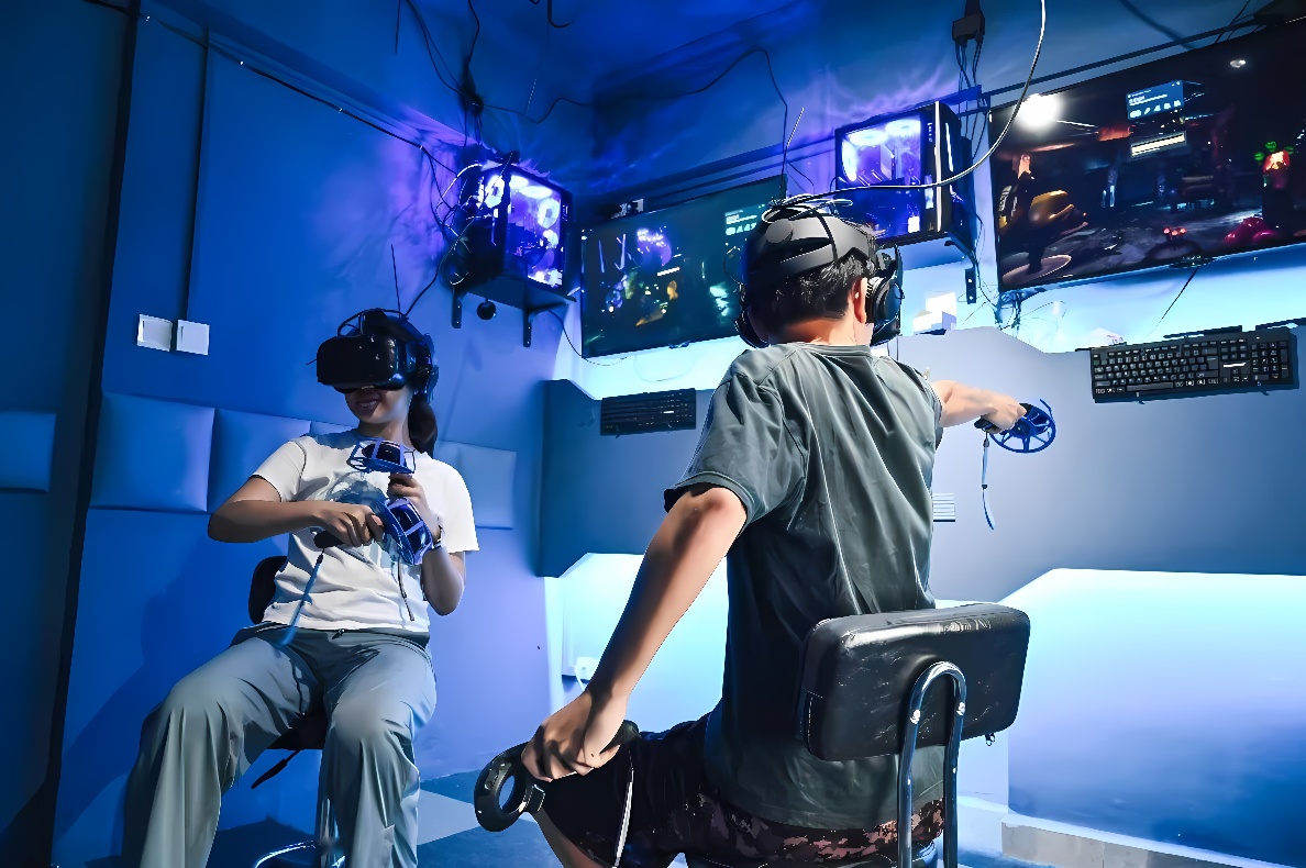 两人戴着VR头盔坐在房间里体验虚拟现实游戏，周围有多个屏幕和电脑设备，氛围科技感十足。