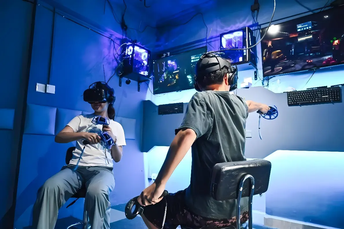 两人戴着虚拟现实头盔坐在房间内，手持控制器，似乎正在体验VR游戏。周围有电脑屏幕和蓝色灯光。