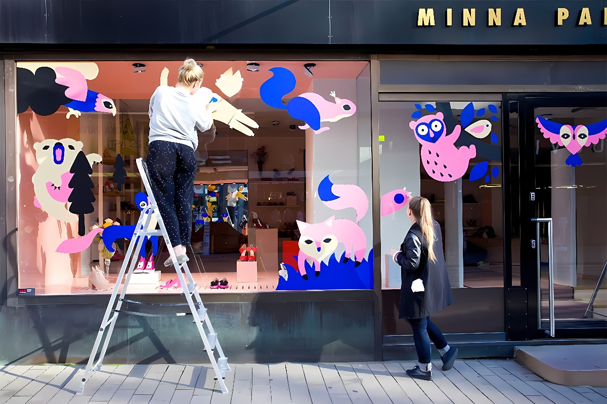 一位女士站在梯子上为商店橱窗贴上色彩鲜艳的卡通动物图案，旁边有位顾客在观看。商店内展示着时尚商品。