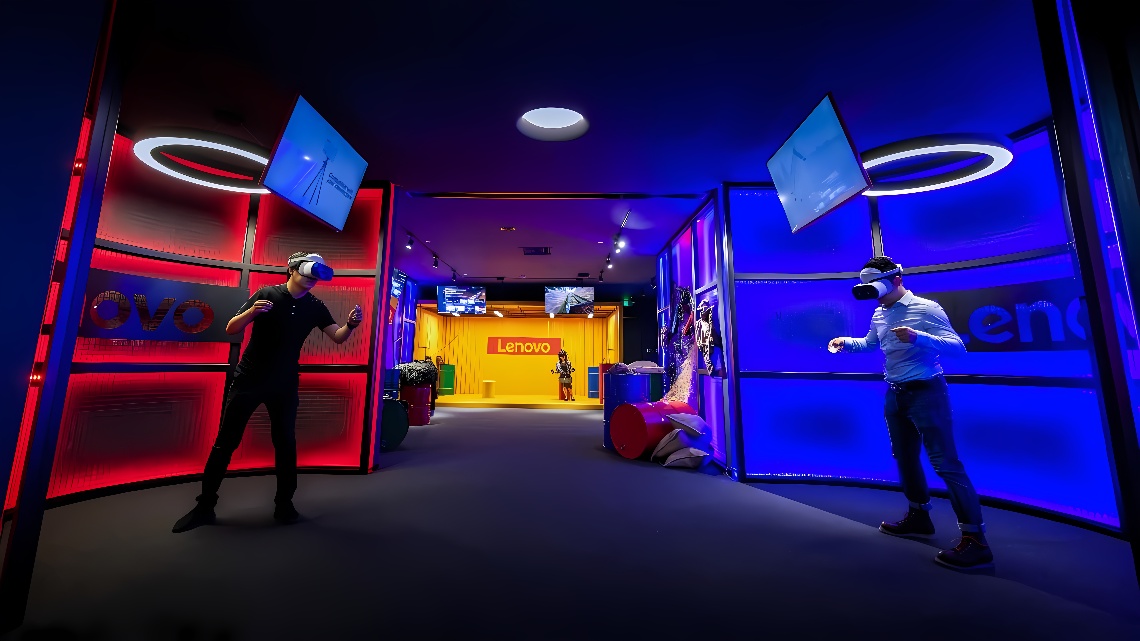 图片展示了两人佩戴虚拟现实头盔，在一个带有Lenovo标志的室内空间进行互动体验，周围环境色彩鲜明，科技感强烈。