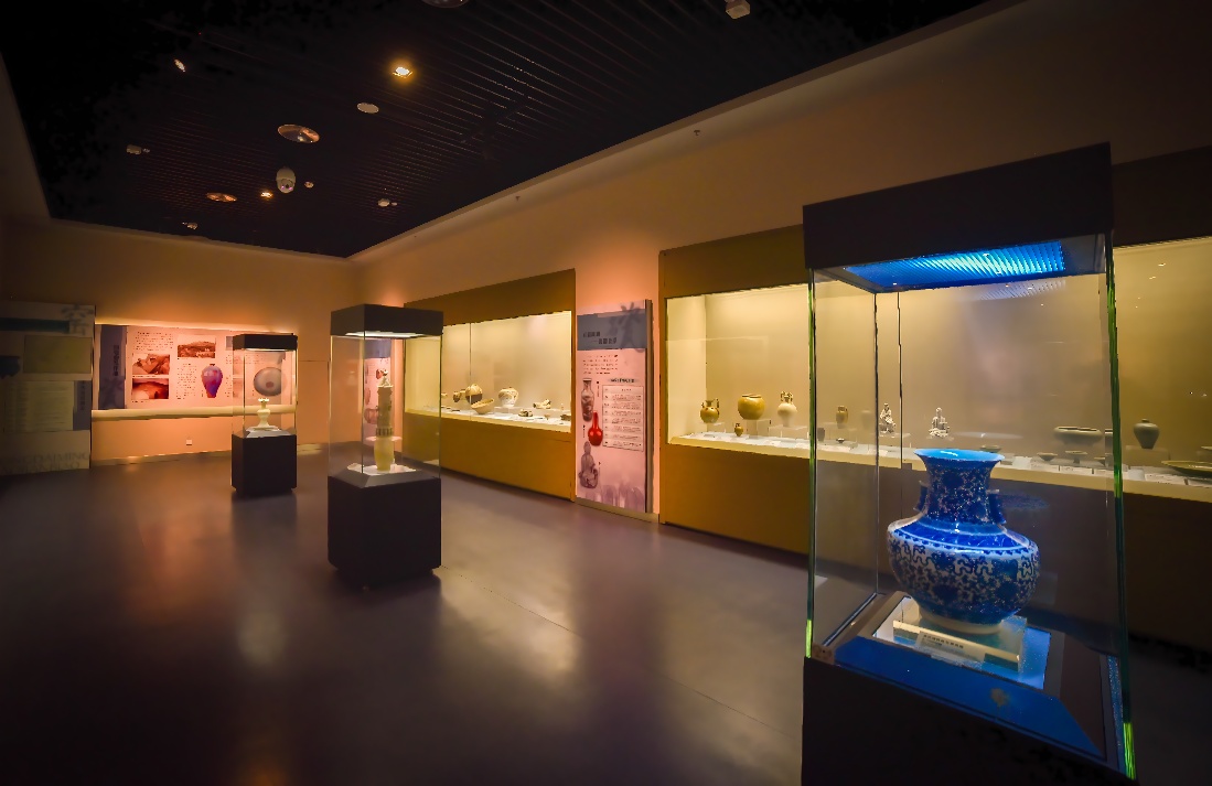 这张图片展示了一个昏暗照明的博物馆内部，里面有几个展示柜，展示着不同的陶瓷艺术品和解说板。