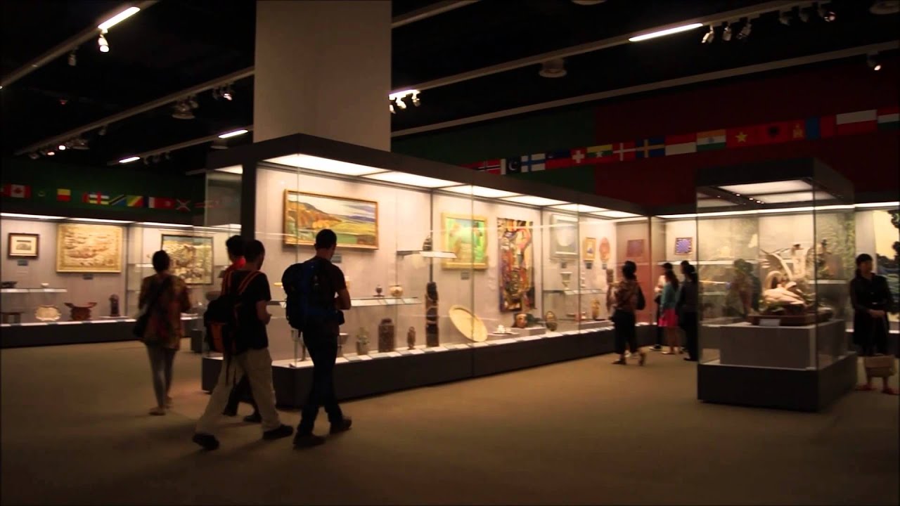 图片展示了一个博物馆内部，参观者正在观看各种展品，包括艺术画作和文物。墙上悬挂着不同国家的旗帜。