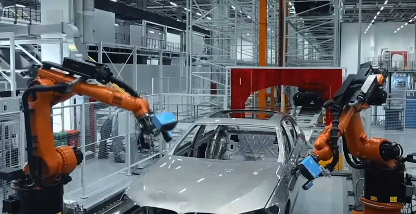 这是一张工业生产线的照片，展示了两个橙色的机械臂在进行汽车装配工作，背景是现代化的工厂环境。