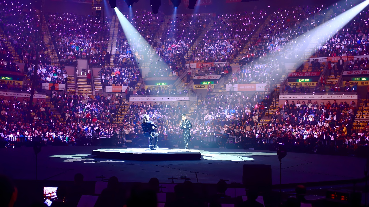 图片展示了一场室内音乐会，舞台中央有艺人表演，现场灯光照射下，观众席满是热情的观众。