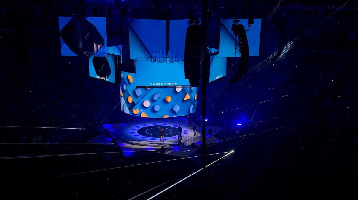 这是一张室内演唱会照片，舞台中央有人表演，四周观众席蓝色灯光映衬，舞台设计现代，悬挂大屏幕和灯光效果。