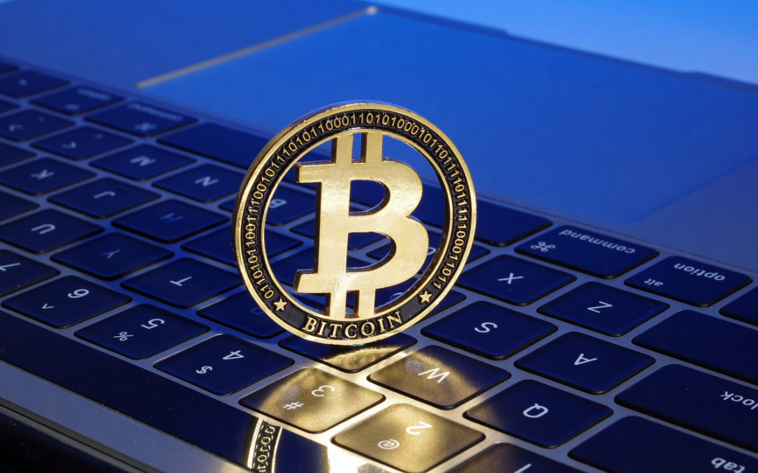这是一枚代表比特币的金色硬币，放置在笔记本电脑键盘上，象征数字货币与现代科技的结合。