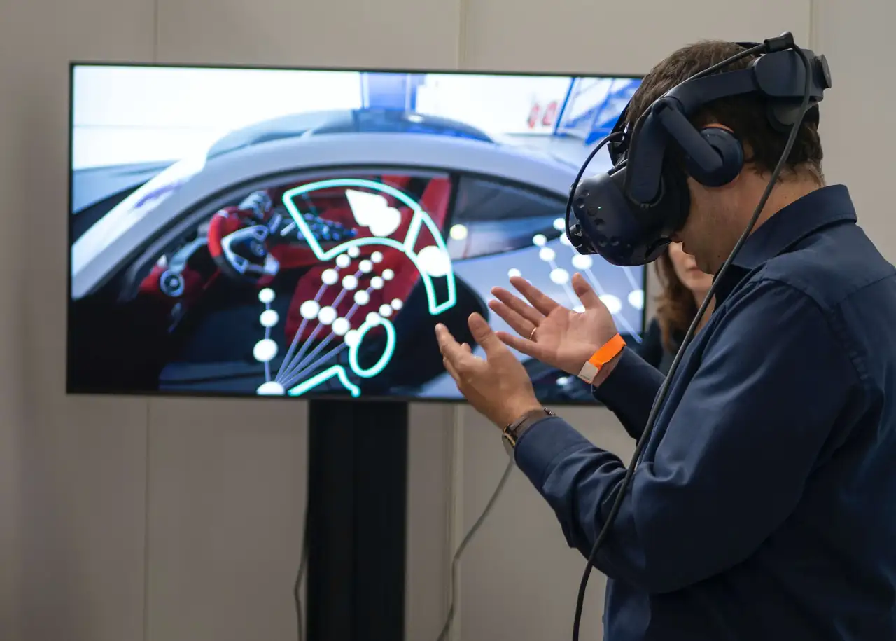 图片展示一位佩戴虚拟现实头盔的男士在体验模拟驾驶场景，他伸出手触摸虚拟图像，背景为大屏幕显示的汽车内饰。