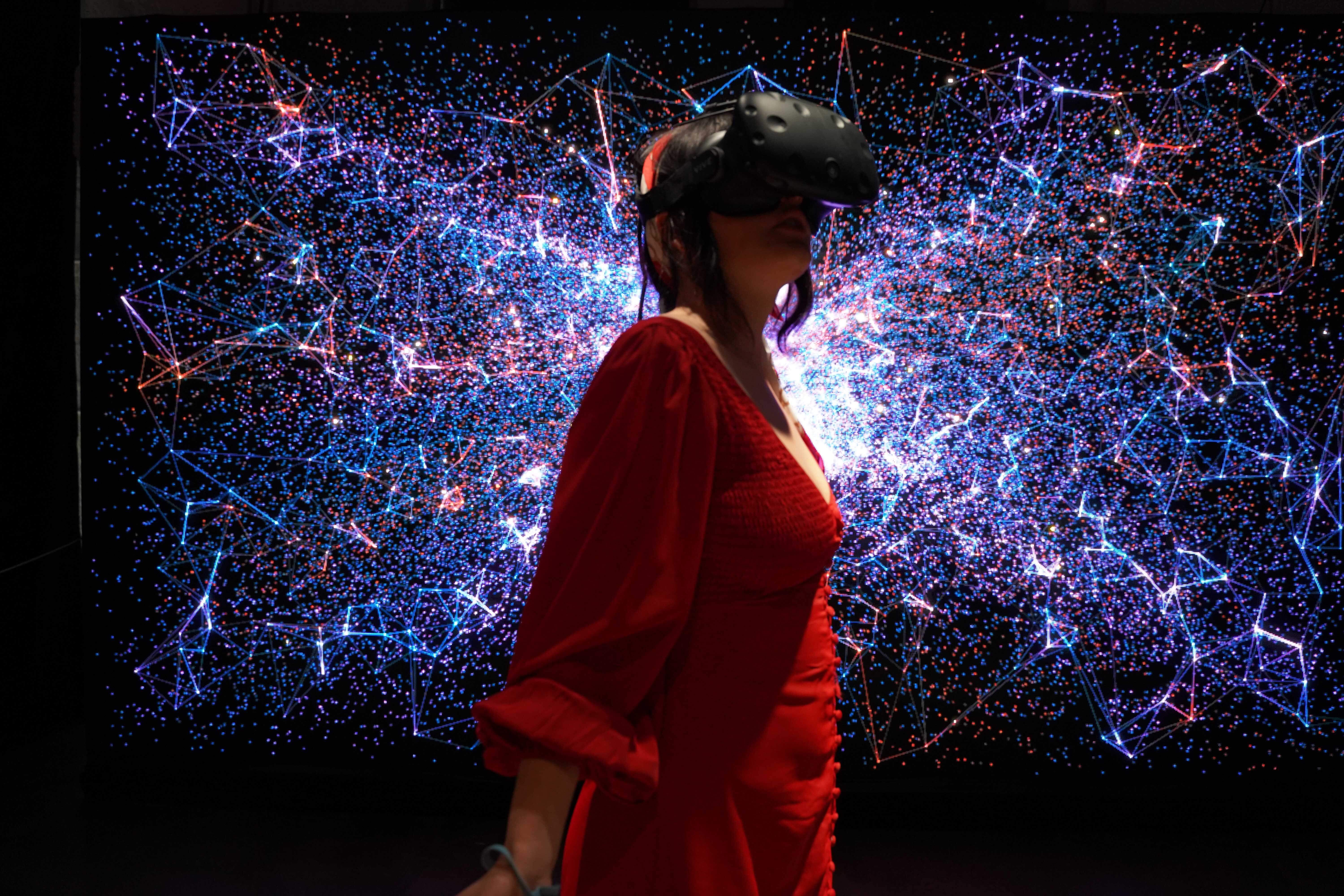 一位女士穿着红色衣服，戴着虚拟现实头盔，站在充满星系和连接线条的虚拟空间前，似乎正在体验沉浸式科技展览。