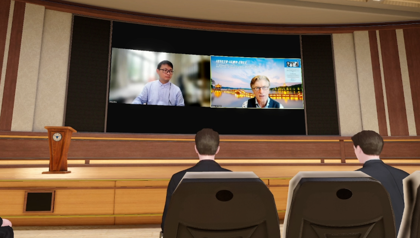 图片展示了一个会议室内部，两个人坐着面向大屏幕，屏幕上正在进行视频会议，有两位参与者在交流。