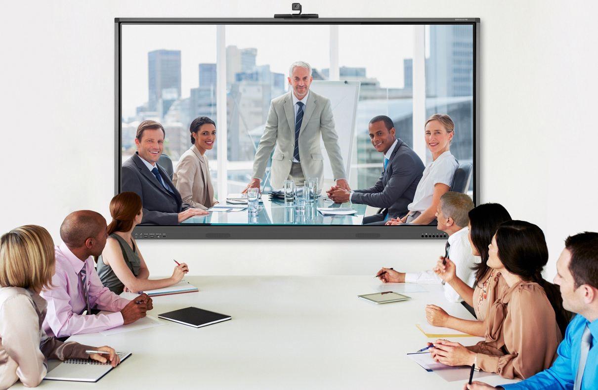 图片展示了一群正装商务人士在会议室内开会，前方有大屏幕显示一位站立的男士，似乎在进行视频会议。
