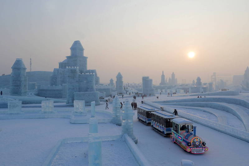 图片展示了一处雪地景区，有冰雕建筑、游客和一辆彩色小火车，天空中太阳微弱，营造出寒冷而宁静的氛围。