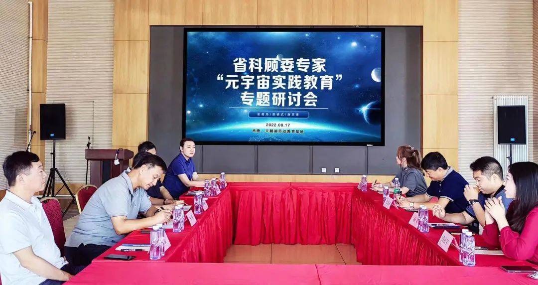 图片展示了一场室内会议，参与者围坐在长会议桌旁，前方是展示着文字内容的大屏幕。