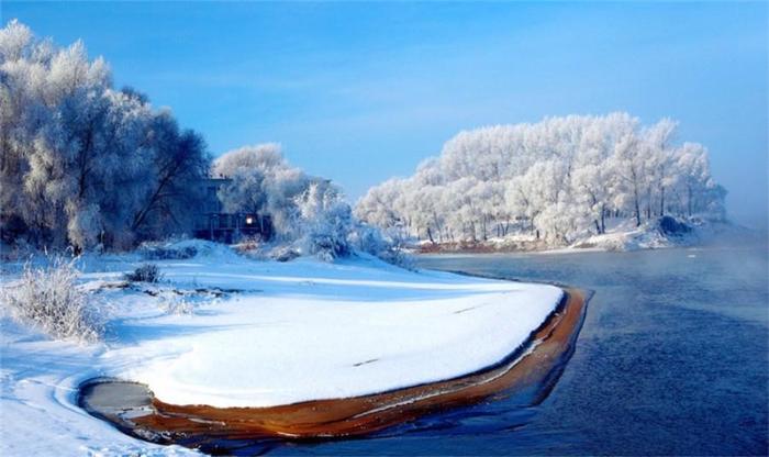 这是一张冬季河景照片。河岸覆盖着雪，河面有薄雾，两岸树木挂满霜花，远处有建筑物，整体景色宁静而美丽。