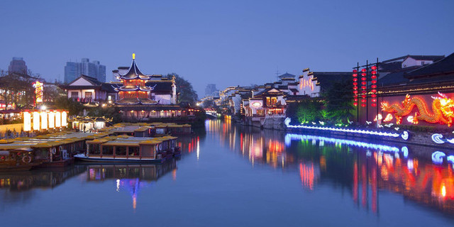 这是一张展示中国传统建筑和现代城市融合的夜景照片，有古典亭台、彩灯、游船和远处现代建筑群。