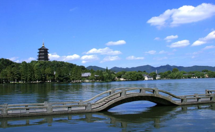 这是一张风景照片，展示了一座古典塔楼，一座拱形桥横跨湖面，背景是蓝天白云和远山。