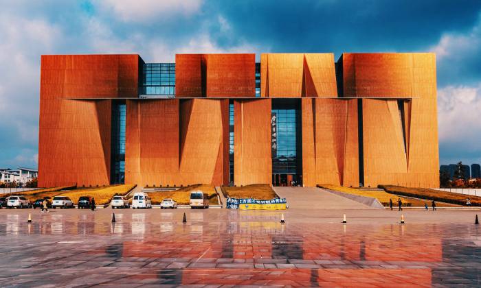 图片展示了一座现代建筑，外观呈现锈红色，有独特的几何形状设计，前方有宽阔的广场和停放的车辆。
