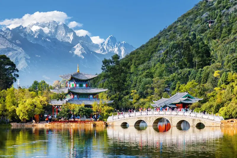 图片展示了一座风格古典的中国桥梁，背后是青翠的山峰和蓝天，显得宁静而美丽。桥上有几位游客正在欣赏风景。