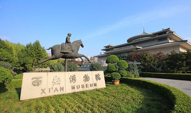 图片展示了西安博物馆前的户外景观，有一座骑马雕塑和博物馆的标识石碑，背后是博物馆的建筑和蓝天。